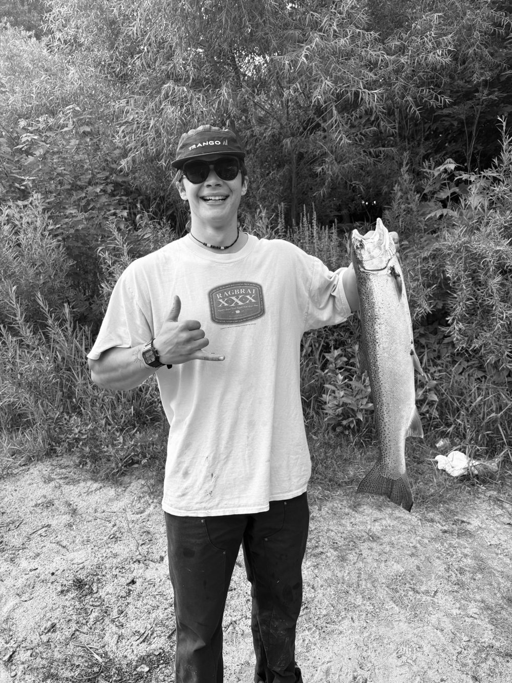 Bridger Hoogeveen, smiling in a cap and sunglasses, holds up a large fish and flashes a shaka sign, shot in black and white
