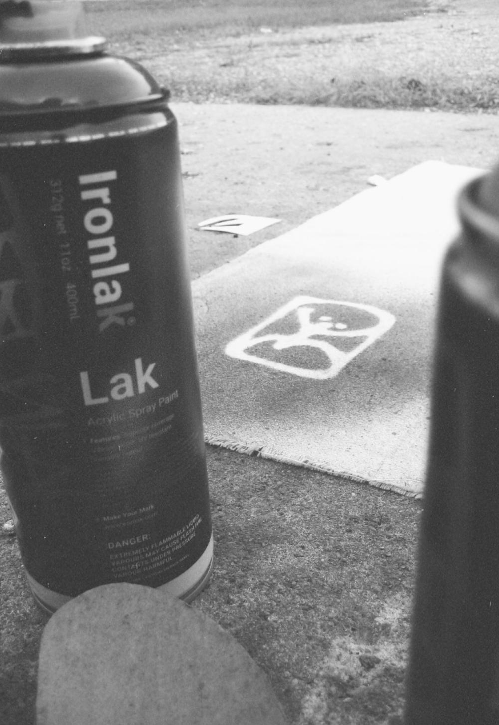 The Dirtbag figure mark sprayed as a white stencil on grey asphalt, with spray cans beside it